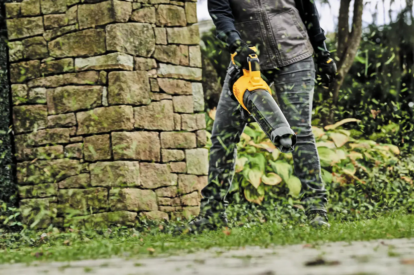 Person using a leaf blower against a stone wall and greenery